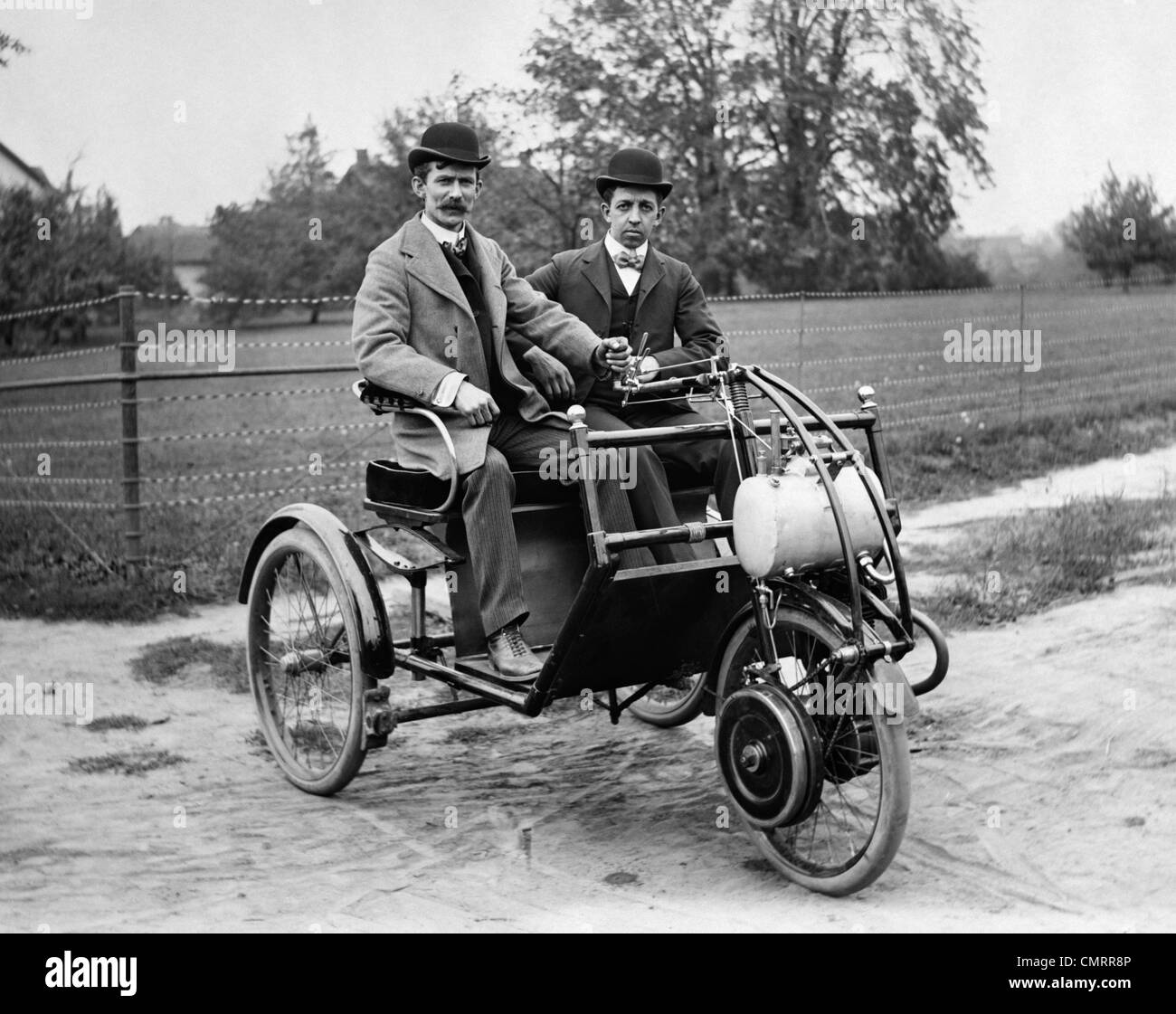1900s TWO MEN IN BOWLER HATS SITTING IN THREE WHEEL MOTORIZED HORSELESS CARRIAGE EARLY AUTOMOBILE WITH TILLER STEERING Stock Photo