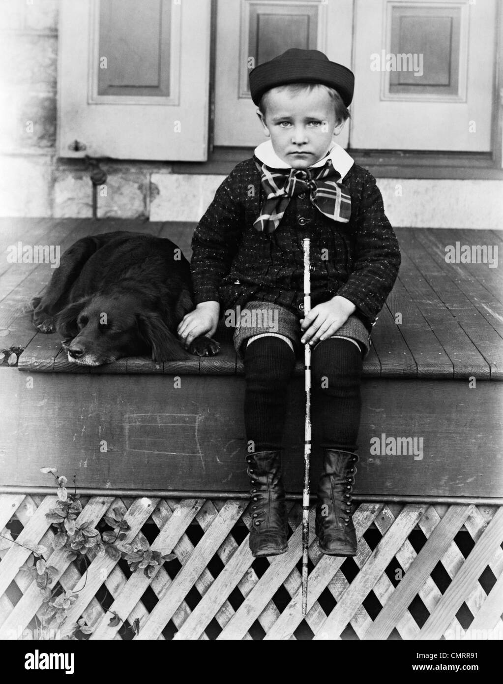 1800s 1890s 1900s TURN OF THE CENTURY SAD BOY HOLDING STICK SITTING ON PORCH NEXT TO DOG Stock Photo