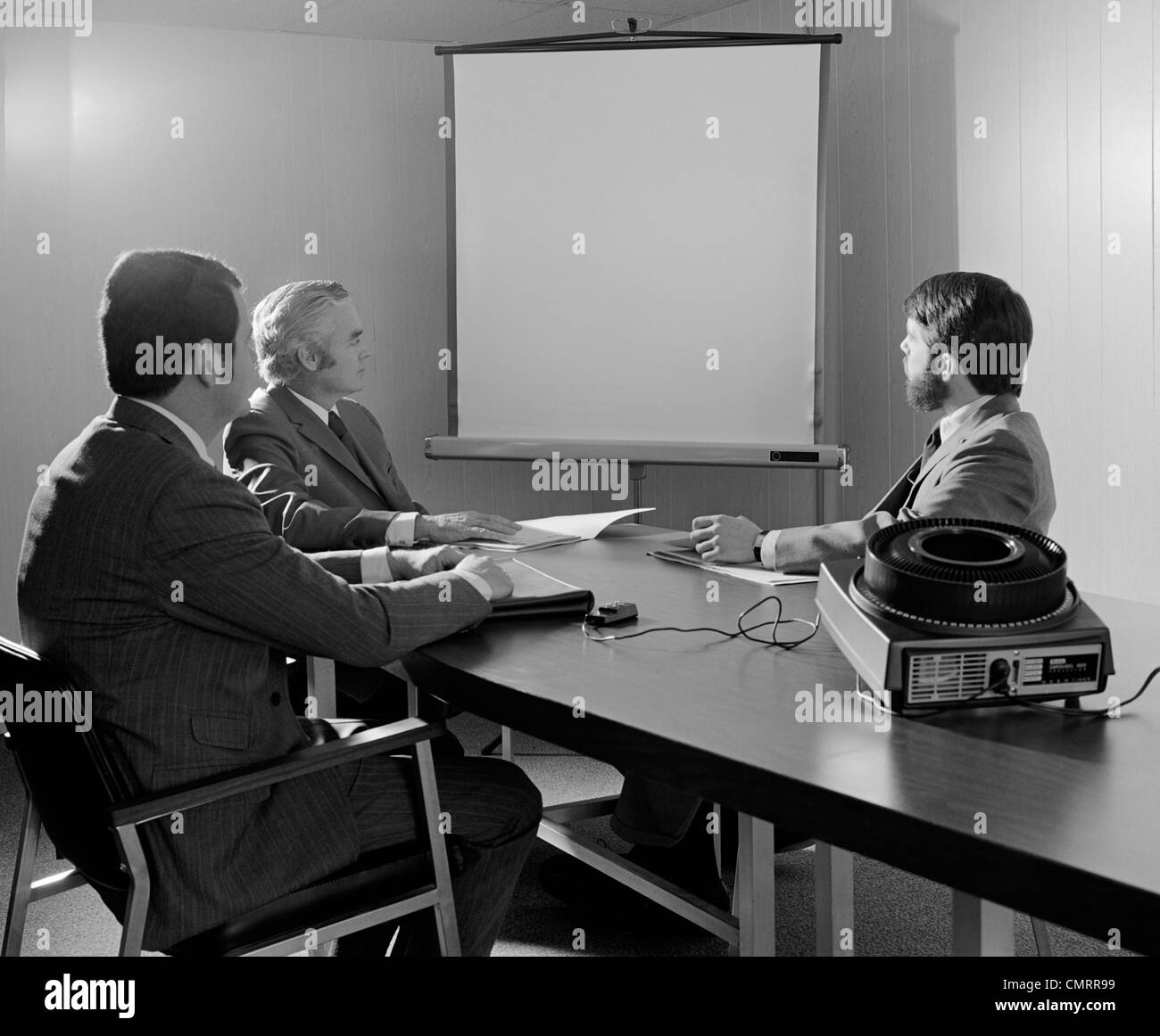 1970s THREE EXECUTIVE BUSINESSMEN CONFERENCE TABLE AUDIOVISUAL PRESENTATION Stock Photo