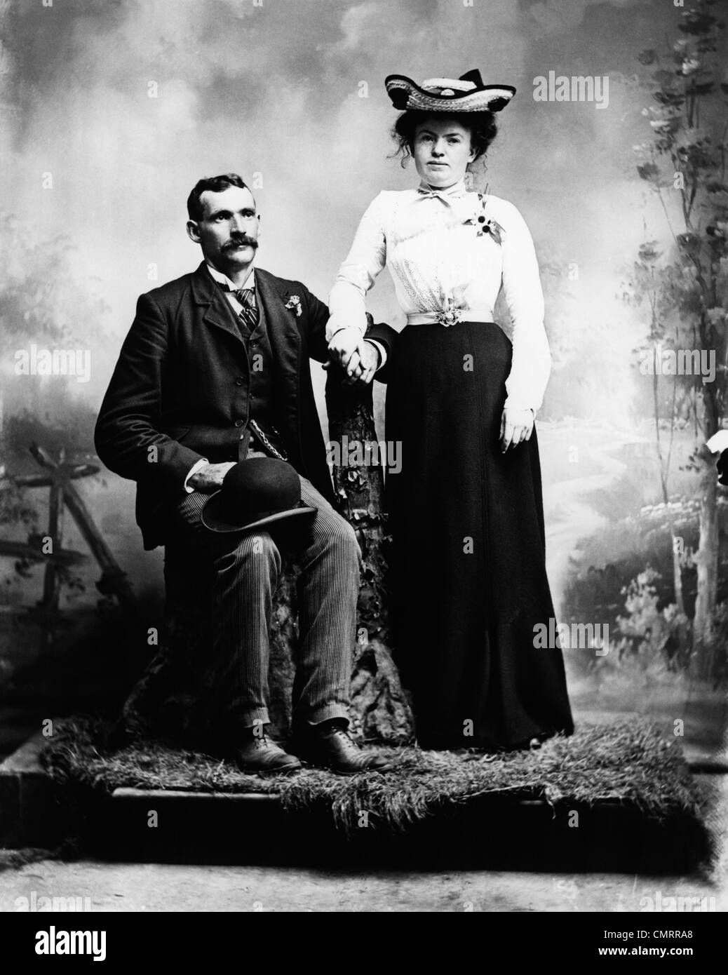 1890s 1900 TURN OF THE CENTURY PORTRAIT SERIOUS COUPLE HOLDING HANDS TOGETHER IN RUSTIC STUDIO SETTING Stock Photo