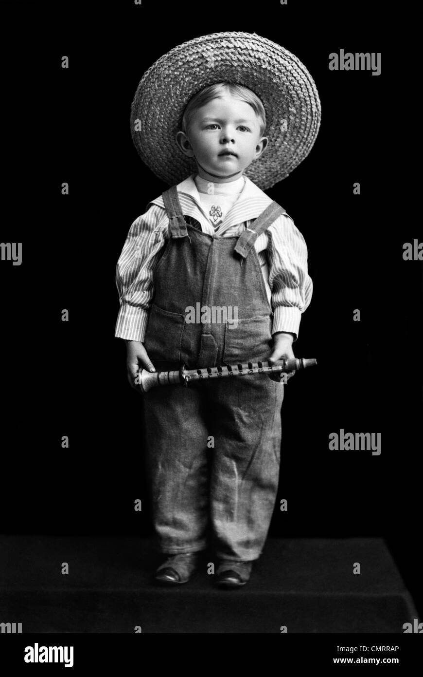 1800s 1890s TURN OF THE CENTURY BOY IN OVERALLS & STRAW HAT HOLDING SMALL CLARINET-LIKE INSTRUMENT Stock Photo