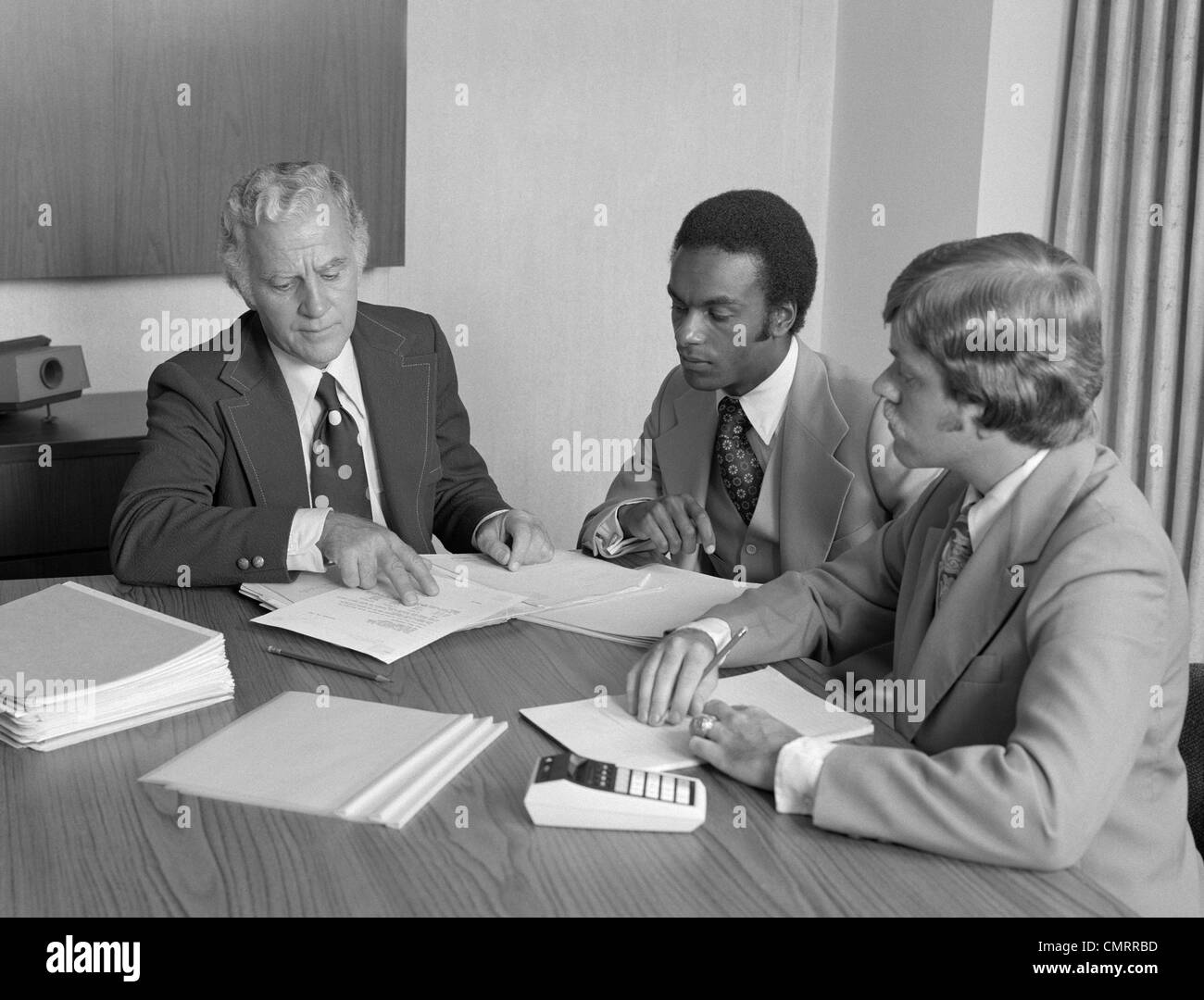 1970s 3 MEN CAUCASIAN & AFRICAN-AMERICAN BUSINESS MEETING AT CONFERENCE TABLE Stock Photo