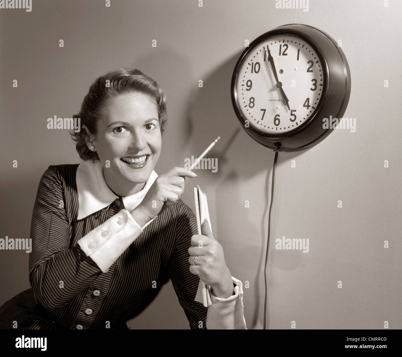 1950s SMILING WOMAN STENOGRAPHER OFFICE WORKER HOLDING STENO PAD POINTING WITH PENCIL TO CLOCK 5 MINUTES TILL QUITTING TIME Stock Photo