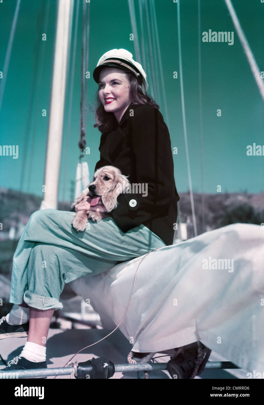 1940s 1950s SMILING WOMAN WEARING SAILING YACHTING OUTFIT SITTING ON EDGE OF SAILBOAT HOLDING PUPPY IN LAP Stock Photo