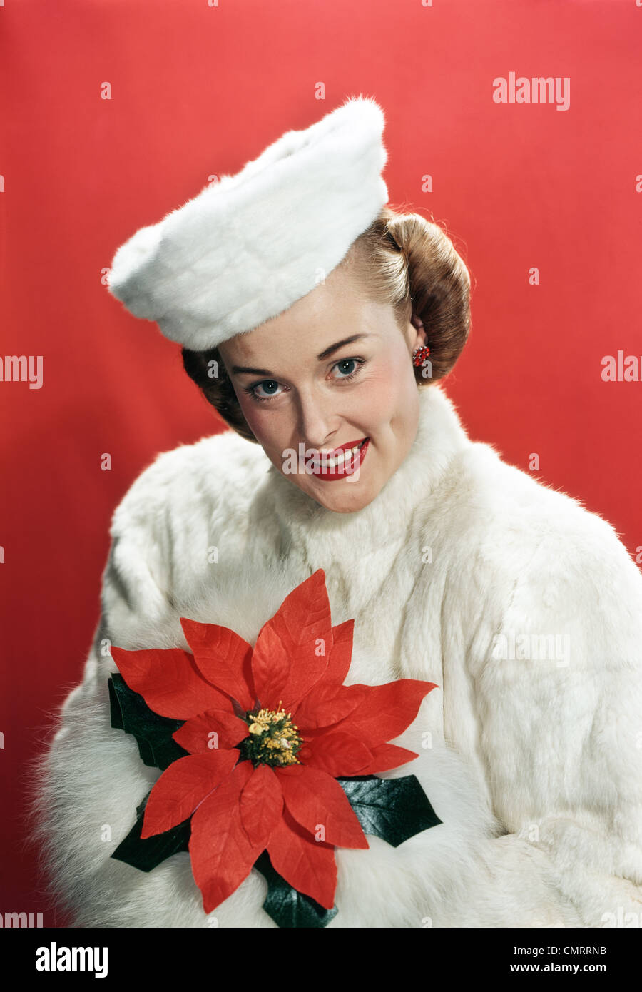 1940s PORTRAIT SMILING WOMAN WEARING WHITE FUR OVERCOAT AND HAT HOLDING POINSETTIA LOOKING AT CAMERA Stock Photo