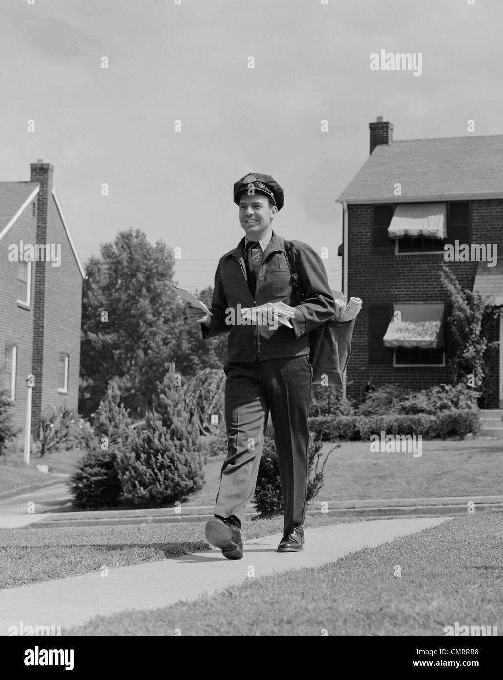 1950s POSTAL MAILMAN WALKING SUBURBAN STREET WEARING UNIFORM DELIVERING MAIL Stock Photo