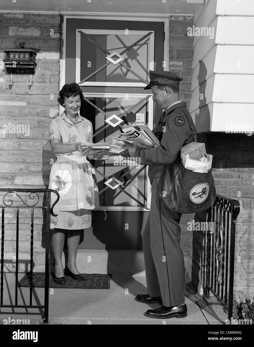 1960s MAN MAILMAN DELIVERING MAIL TO WOMAN HOUSEWIFE IN FRONT OF SUBURBAN HOUSE OUTDOOR Stock Photo