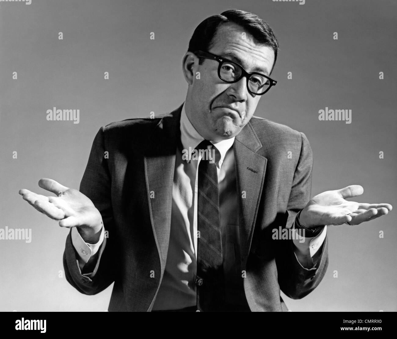 1960s BUSINESSMAN SHRUGGING SHOULDERS LOOKING AT CAMERA HANDS EXTENDED PALMS UP Stock Photo