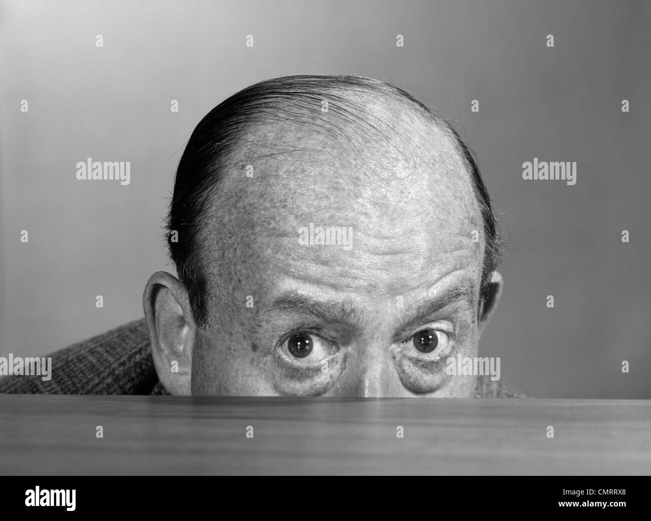 1960s BALDING MAN PEEKING OVER DESKTOP WITH ONLY TOP HALF OF HEAD SHOWING LOOKING AT CAMERA Stock Photo