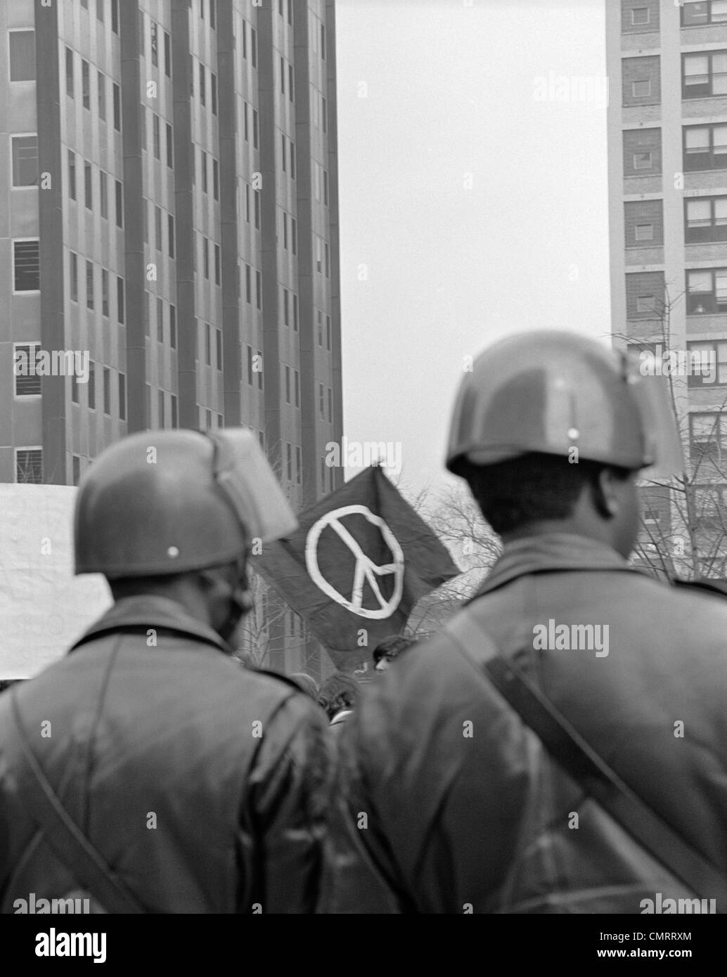 1970s BACK VIEW OF TWO POLICEMAN IN HELMETS AT DEMONSTRATION WITH PEACE SIGN IN BACKGROUND FRAMED BETWEEN THEIR HEADS Stock Photo