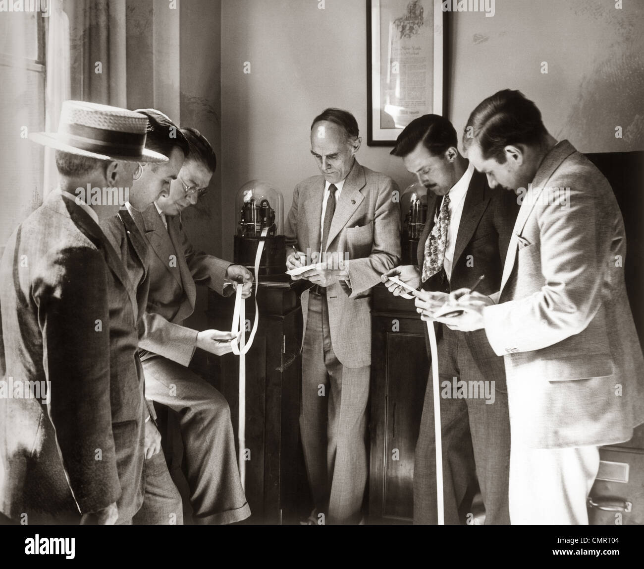 1930s BROKERS BUSINESSMEN READING TICKER TAPE AS IT COMES OUT OF GLASS DOMED MACHINE Stock Photo