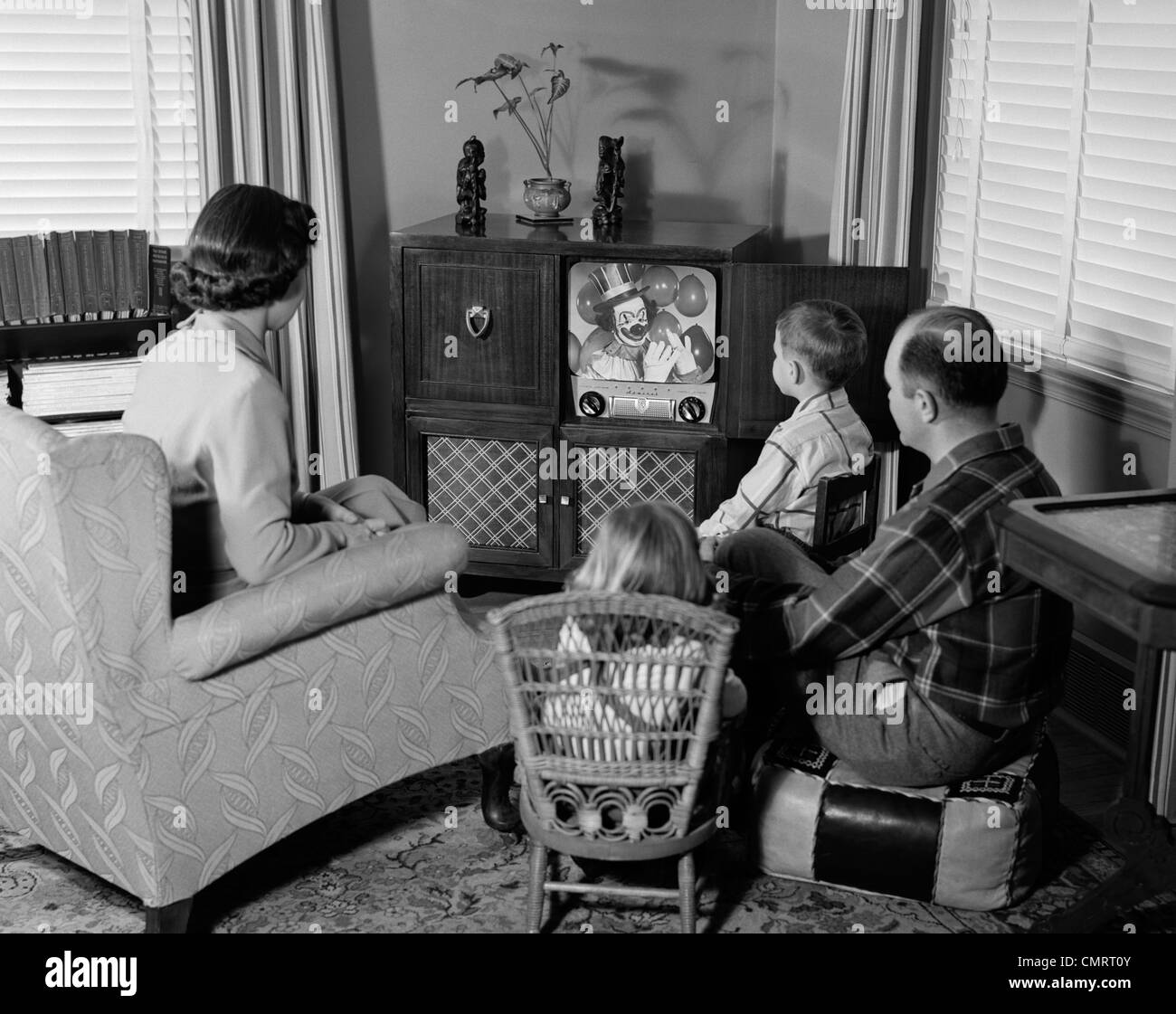 1950s BACK VIEW OF FAMILY OF 4 GATHERED AROUND TV SET WATCHING CLOWN WITH BALLOONS Stock Photo