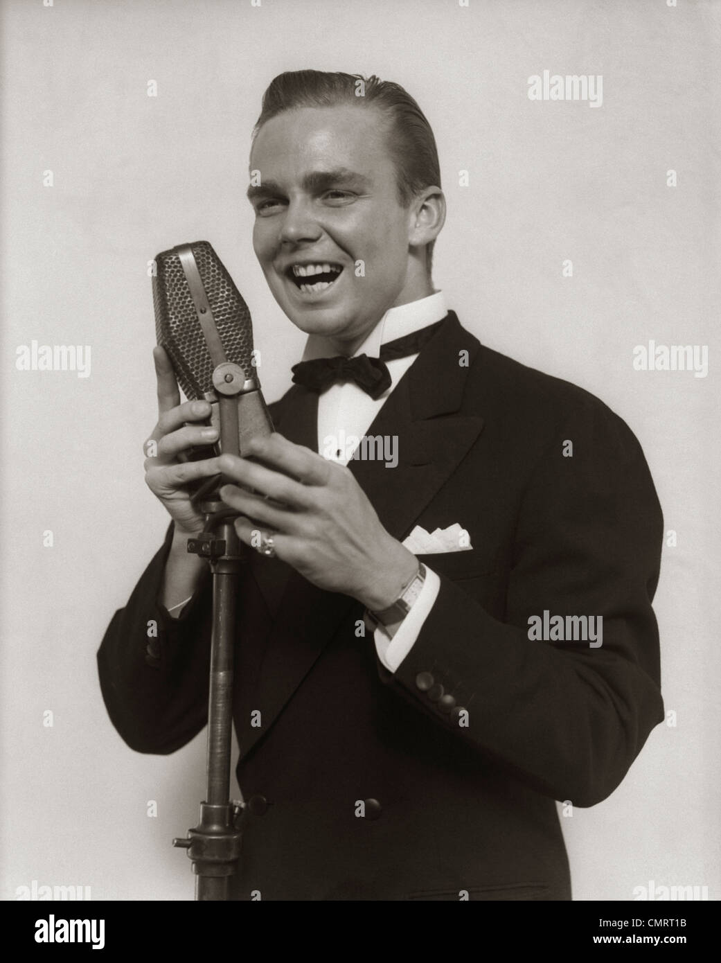 1920s 1930s SMILING MAN RADIO SINGER ENTERTAINER CROONER IN TUXEDO SINGING INTO MICROPHONE Stock Photo