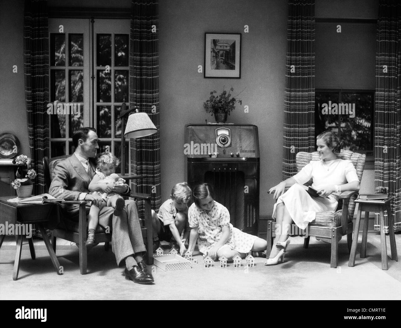 1930s 1940s FAMILY IN LIVING ROOM LISTENING TO RADIO WHILE CHILDREN PLAY WITH TOY HOUSES ON FLOOR Stock Photo