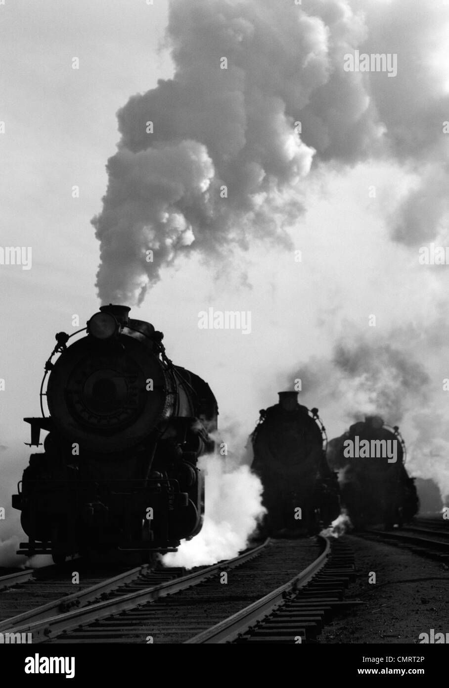 1930s 1940s HEAD-ON VIEW OF THREE STEAM ENGINES SILHOUETTED AGAINST BILLOWING SMOKE AND STEAM OUTDOOR Stock Photo