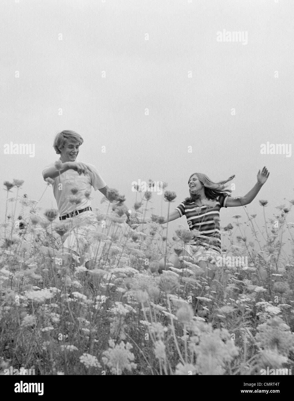 1970s YOUNG COUPLE HOLDING HANDS RUNNING THROUGH FIELD OF FLOWERS Stock Photo