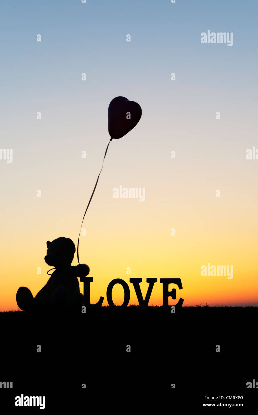 Teddy bear holding a heart shape balloon next too the word LOVE at sunrise Stock Photo