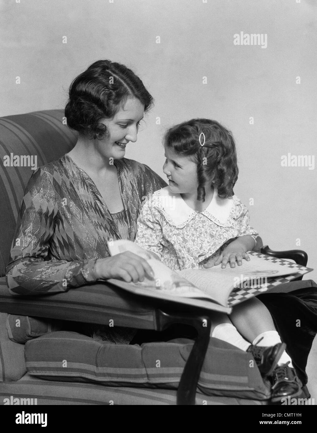 1920s 1930s MOTHER & DAUGHTER SMILING SITTING IN CHAIR READING BOOK Stock Photo