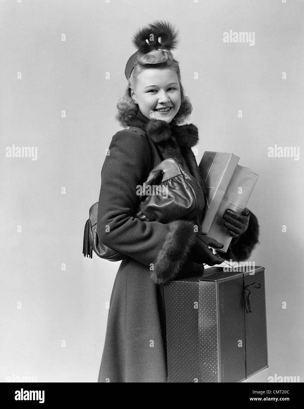 1940s 1950s SMILING YOUNG BLOND WOMAN WEARING HAT FUR TRIMMED COAT CARRYING PACKAGES LOOKING AT CAMERA Stock Photo