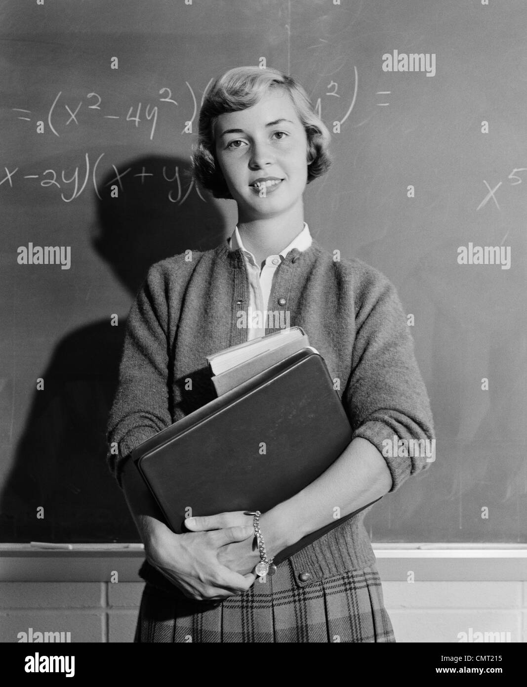 1960s TEENAGE GIRL HOLDING SCHOOL BOOKS STANDING IN FRONT OF BLACKBOARD LOOKING AT CAMERA Stock Photo