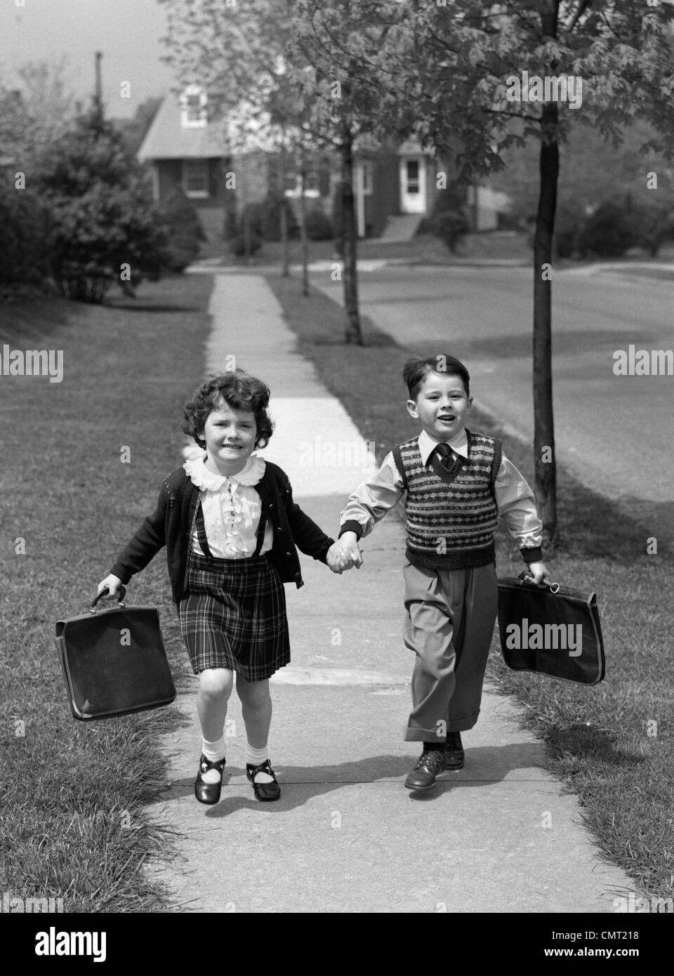 1950s BOY AND GIRL CARRYING BOOK BAGS WALKING TOGETHER HAND IN HAND ON SIDEWALK TO OR FROM SCHOOL OUTDOOR Stock Photo