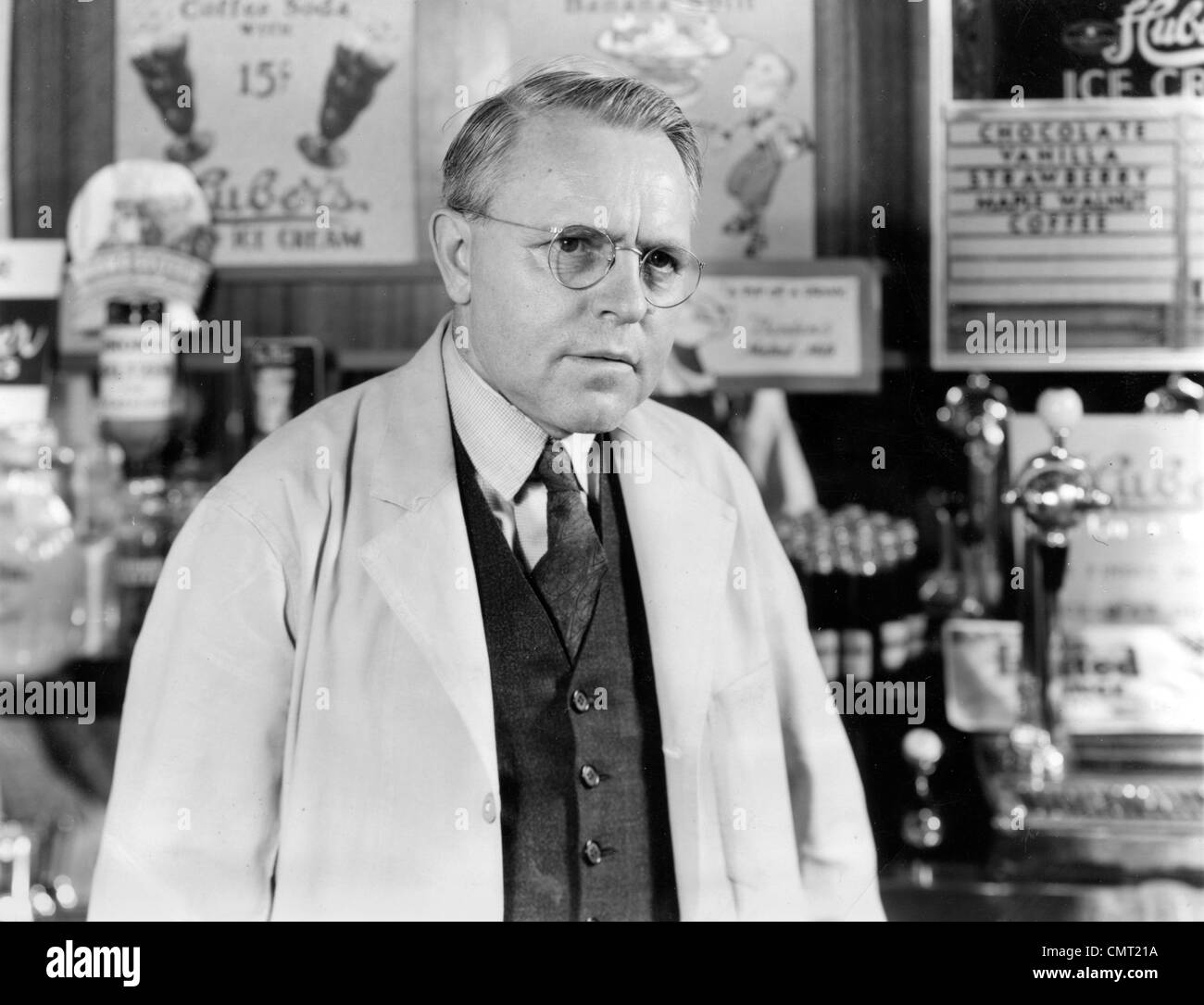 1930s 1940s DRUG STORE SODA FOUNTAIN MAN PHARMACIST WEARING WHITE COAT AND GLASSES LISTENING Stock Photo