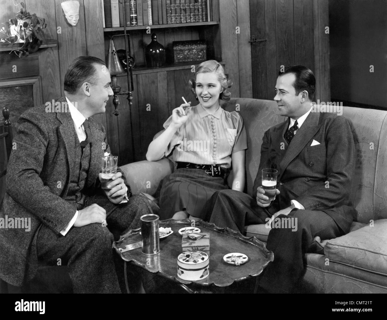 1940s TWO MEN AND ONE WOMAN SOCIAL GROUP SITTING ON COUCH DRINKING BEER SMOKING CIGARETTES TALKING Stock Photo