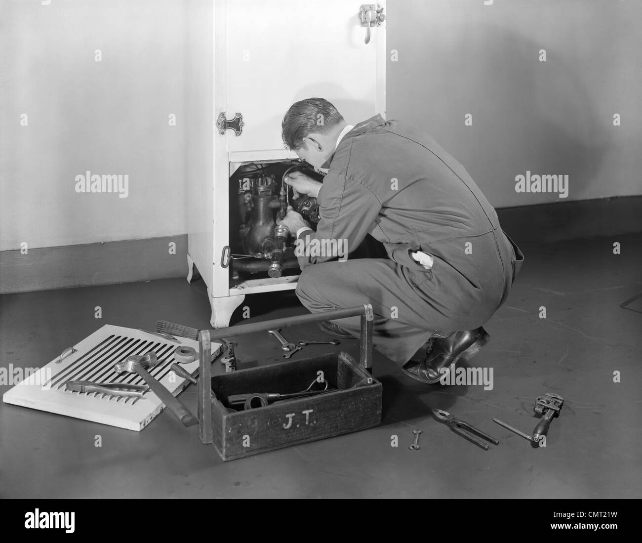 1930s 1940s MAN WORKMAN FIXING REFRIGERATION MACHINERY OF REFRIGERATOR IN KITCHEN REPAIR SERVICE Stock Photo