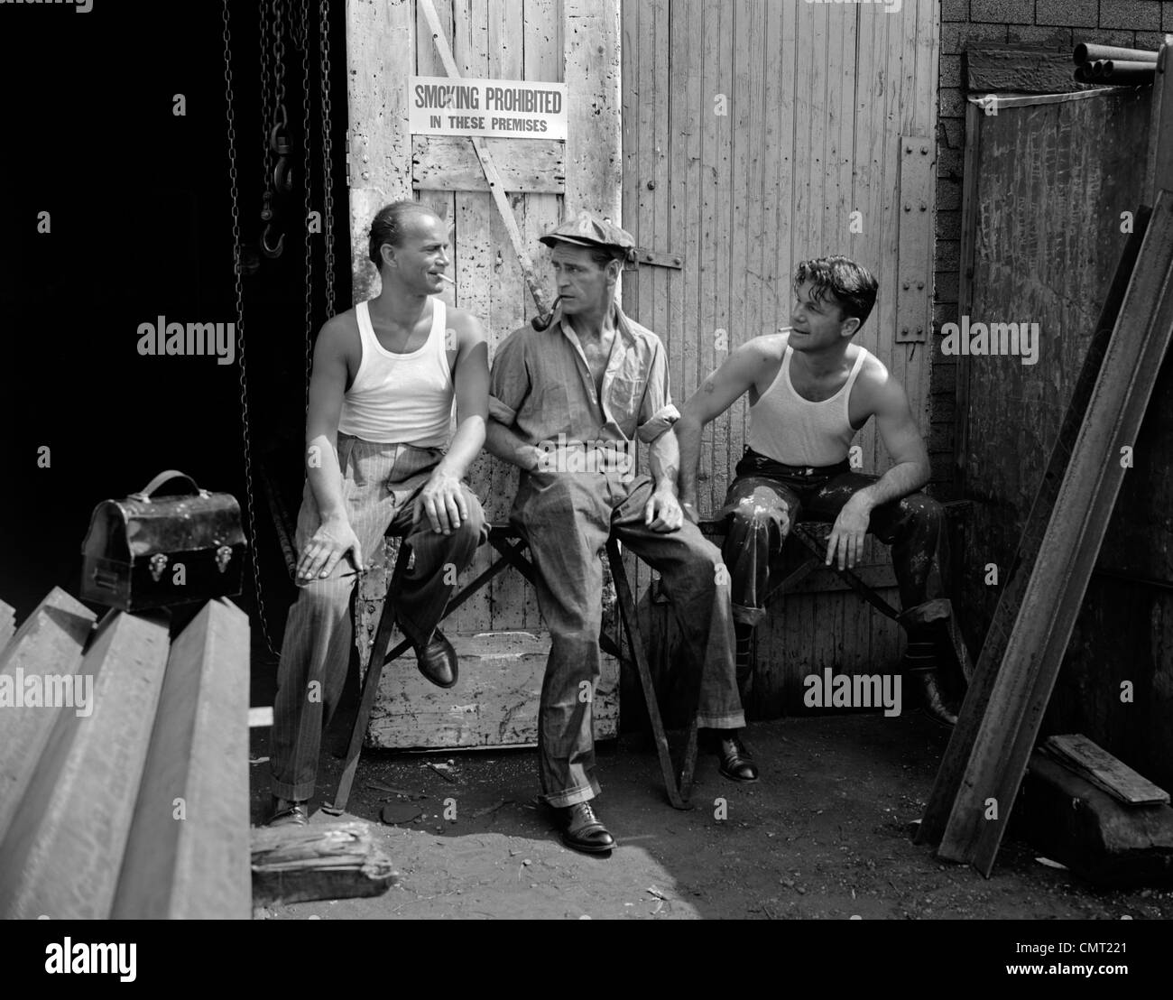 1930s 1940s THREE WORKMEN SITTING UNDER NO SMOKING SIGN SMOKING CIGARETTE AND PIPE JOB SAFETY Stock Photo