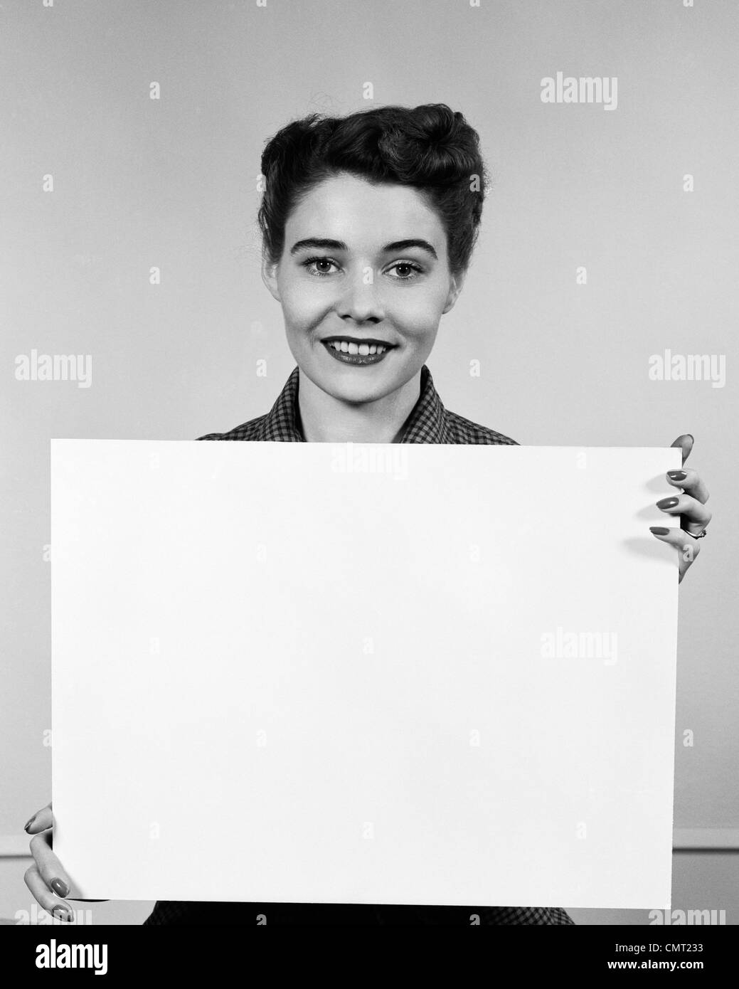 1950s WOMAN HOLDING BLANK SIGN IN FRONT OF CHEST SMILING INDOOR Stock Photo