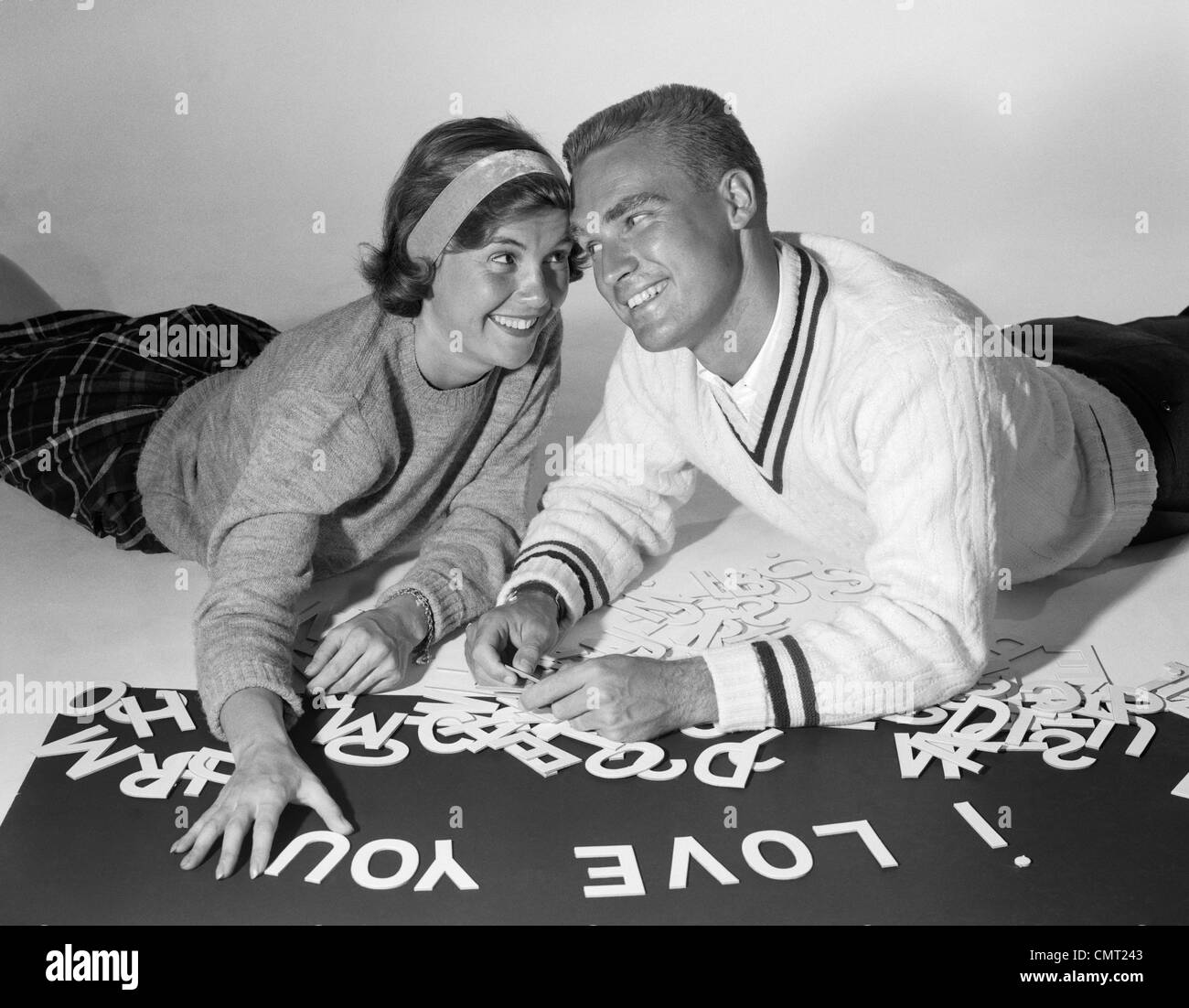 1960s SMILING COLLEGIATE COUPLE LYING ON STOMACHS WITH HEADS PRESSED TOGETHER SPELLING OUT I LOVE YOU WITH CUT-OUT LETTERS Stock Photo