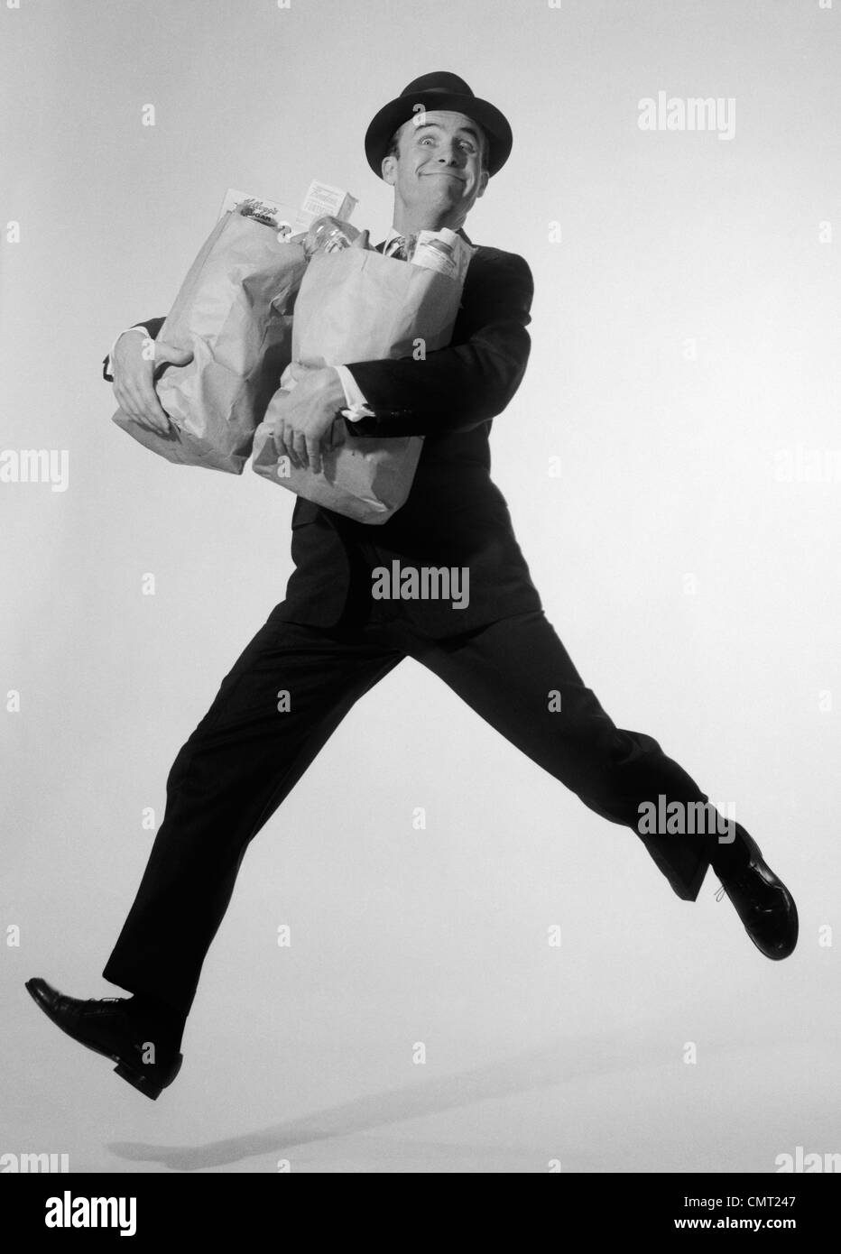 1960s MAN IN SUIT & FEDORA LEAPING FORWARD WITH GROCERY BAGS IN ARMS LOOKING AT CAMERA Stock Photo