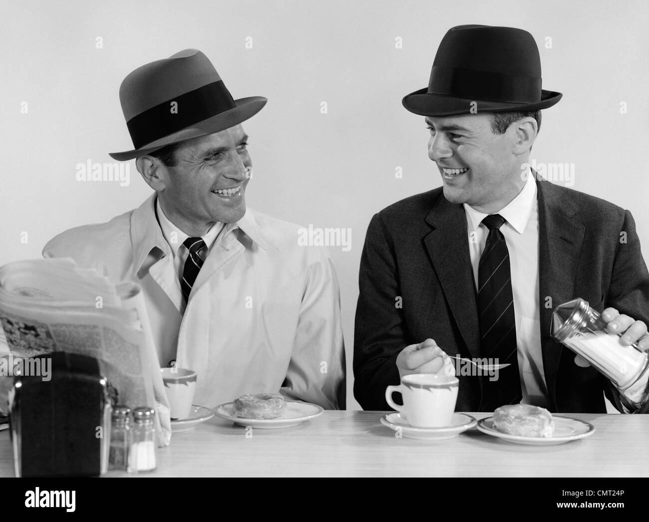 1960s TWO BUSINESSMEN WEARING HATS DRINKING COFFEE SMILING TALKING SITTING TOGETHER AT A DINER LUNCH COUNTER Stock Photo