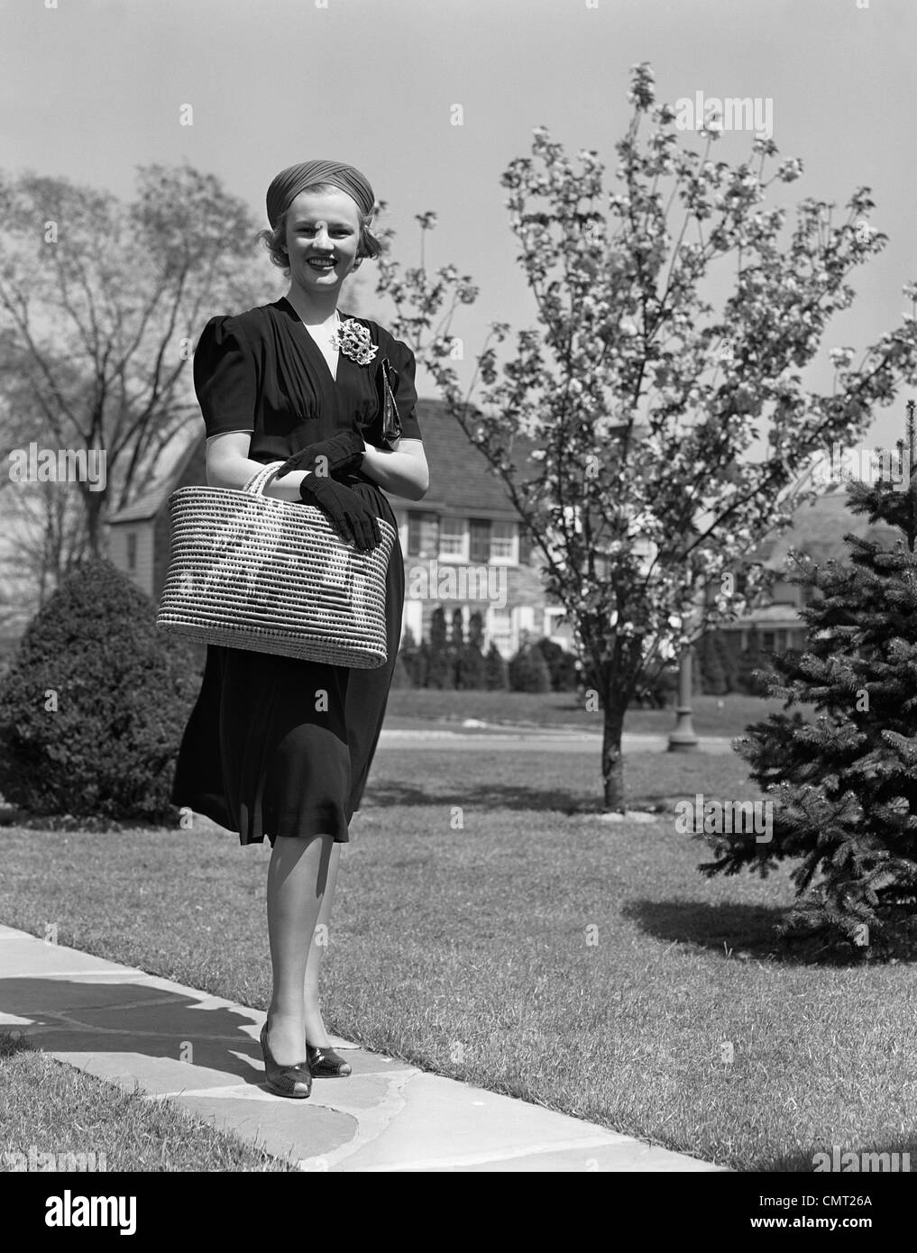 1930s 1940s STYLISHLY DRESSED WOMAN WALKING DOWN SUBURBAN STREET CARRYING STRAW BAG Stock Photo