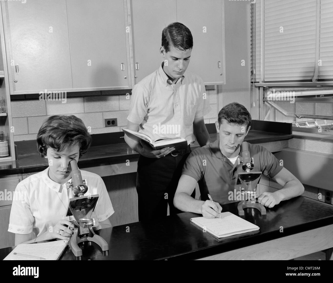 1960s THREE STUDENTS USING MICROSCOPES TWO BOYS ONE GIRL INDOOR HIGH SCHOOL Stock Photo