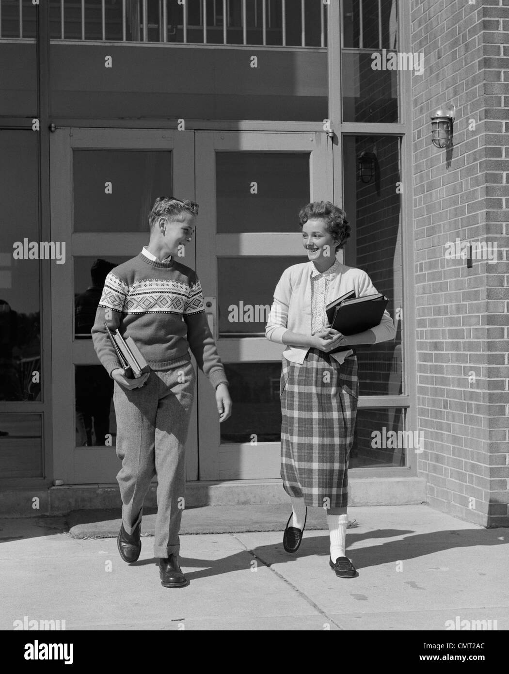 1950s TWO TEENAGE STUDENTS BOY AND GIRL WALKING AWAY FROM SCHOOL DOORS CARRYING BOOKS Stock Photo