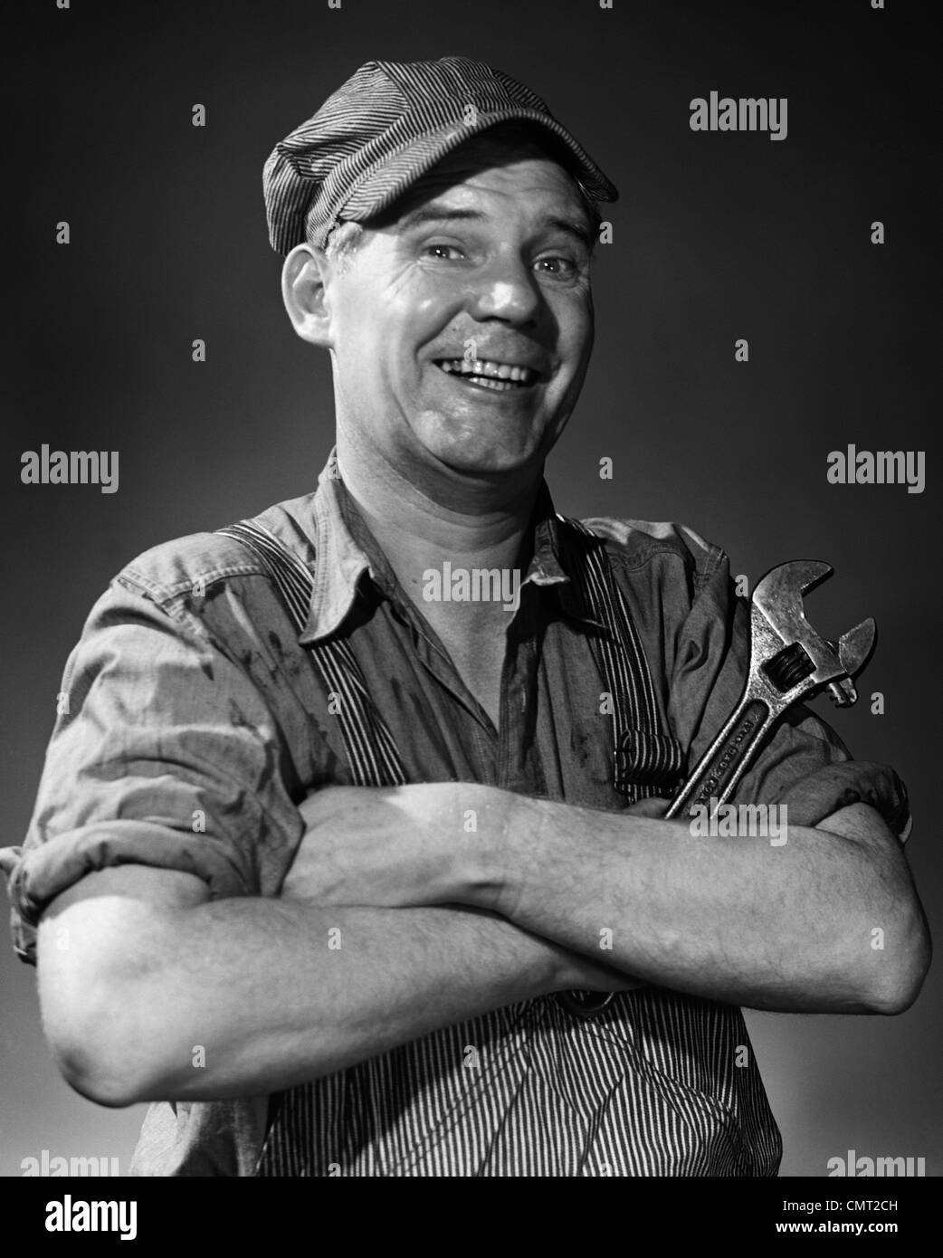 1950s 1960s SMILING PORTRAIT MAN IN STRIPED CAP & OVERALLS CROSSING ARMS HOLDING MONKEY WRENCH LOOKING AT CAMERA Stock Photo