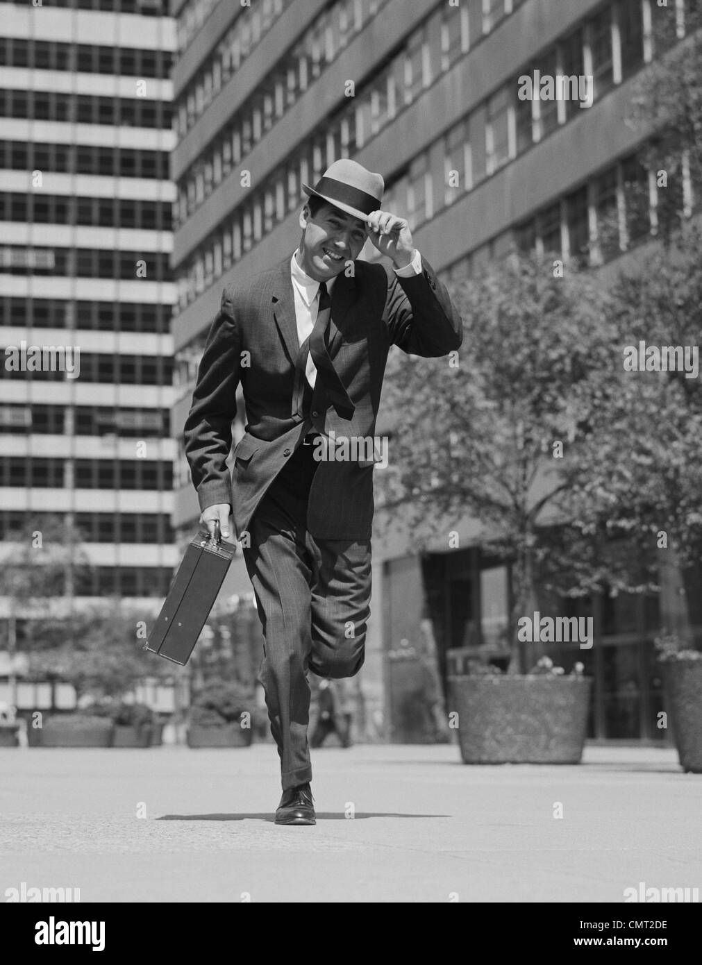 1960s BUSINESSMAN CARRYING BRIEFCASE HOLDING HIS HAT ON RUNNING DOWN URBAN SIDEWALK Stock Photo