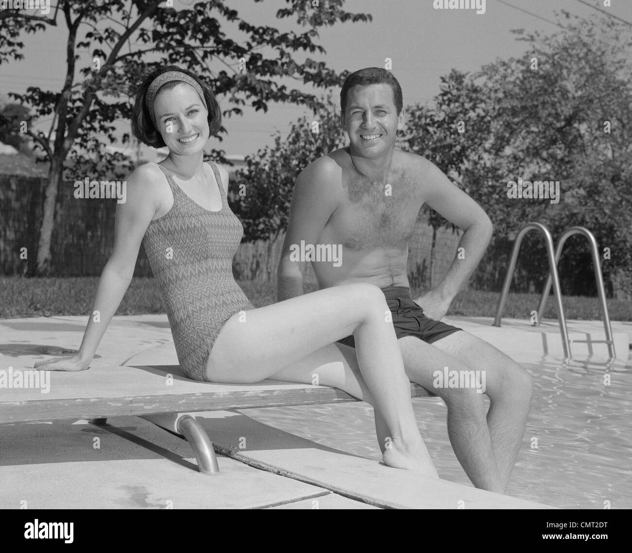 1960s MAN WOMAN COUPLE SITTING ON DIVING BOARD ON SIDE OF SWIMMING POOL Stock Photo