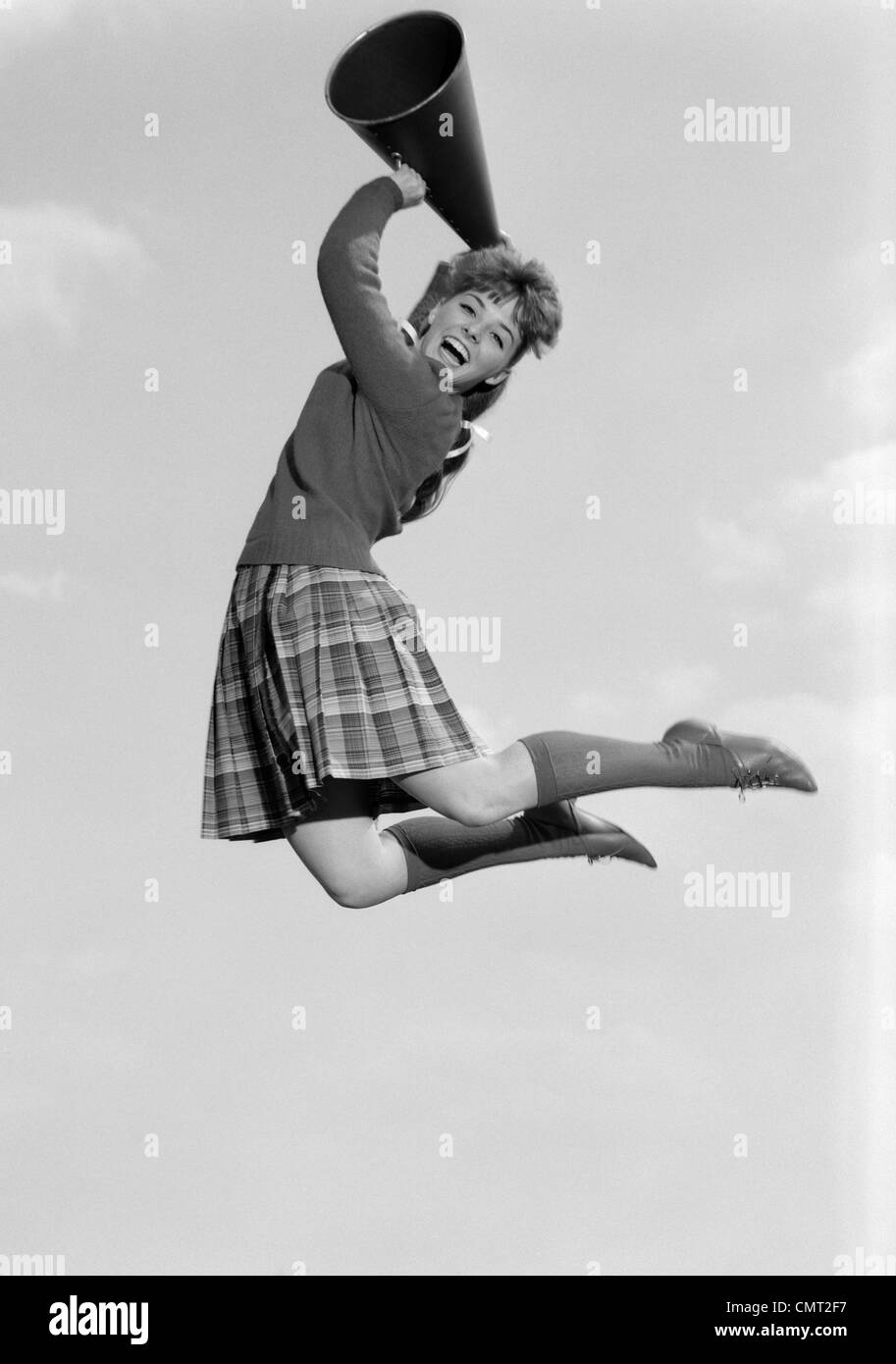 1960s TEENAGE GIRL CHEERLEADER CHEERING JUMPING INTO THE AIR HOLDING MEGAPHONE LOOKING AT CAMERA Stock Photo