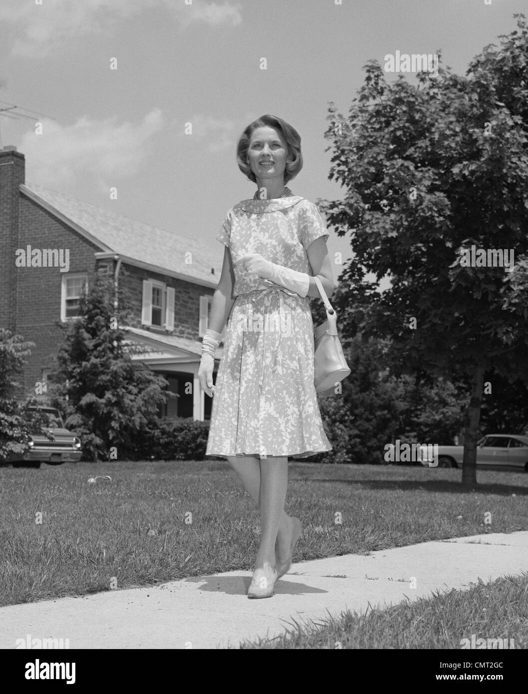 1960s WOMAN WEARING PRINT DRESS HOLDING HANDBAG WALKING DOWN SUBURBAN STREET Stock Photo