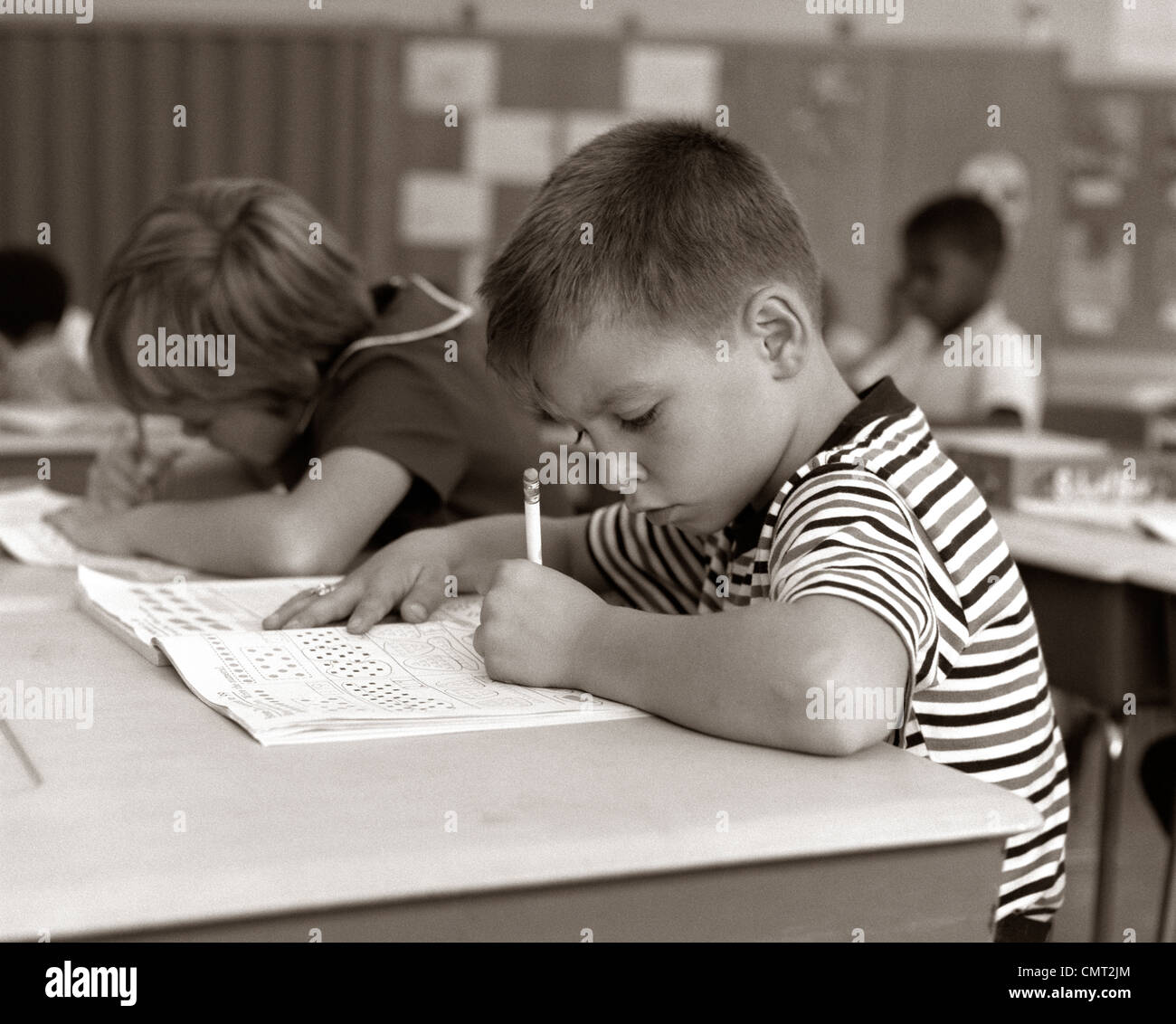 1960s BOY STRIPED T-SHIRT ELEMENTARY SCHOOL CLASSROOM SITTING DESK WRITING TEST Stock Photo