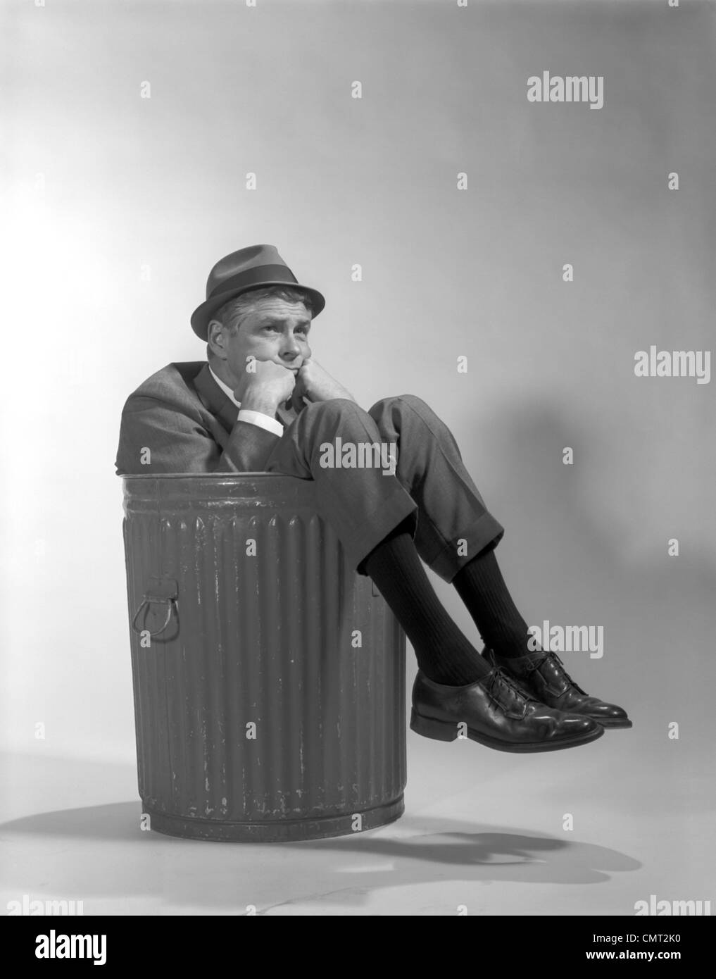 1960s BUSINESSMAN IN SUIT AND HAT SITTING IN TRASHCAN Stock Photo