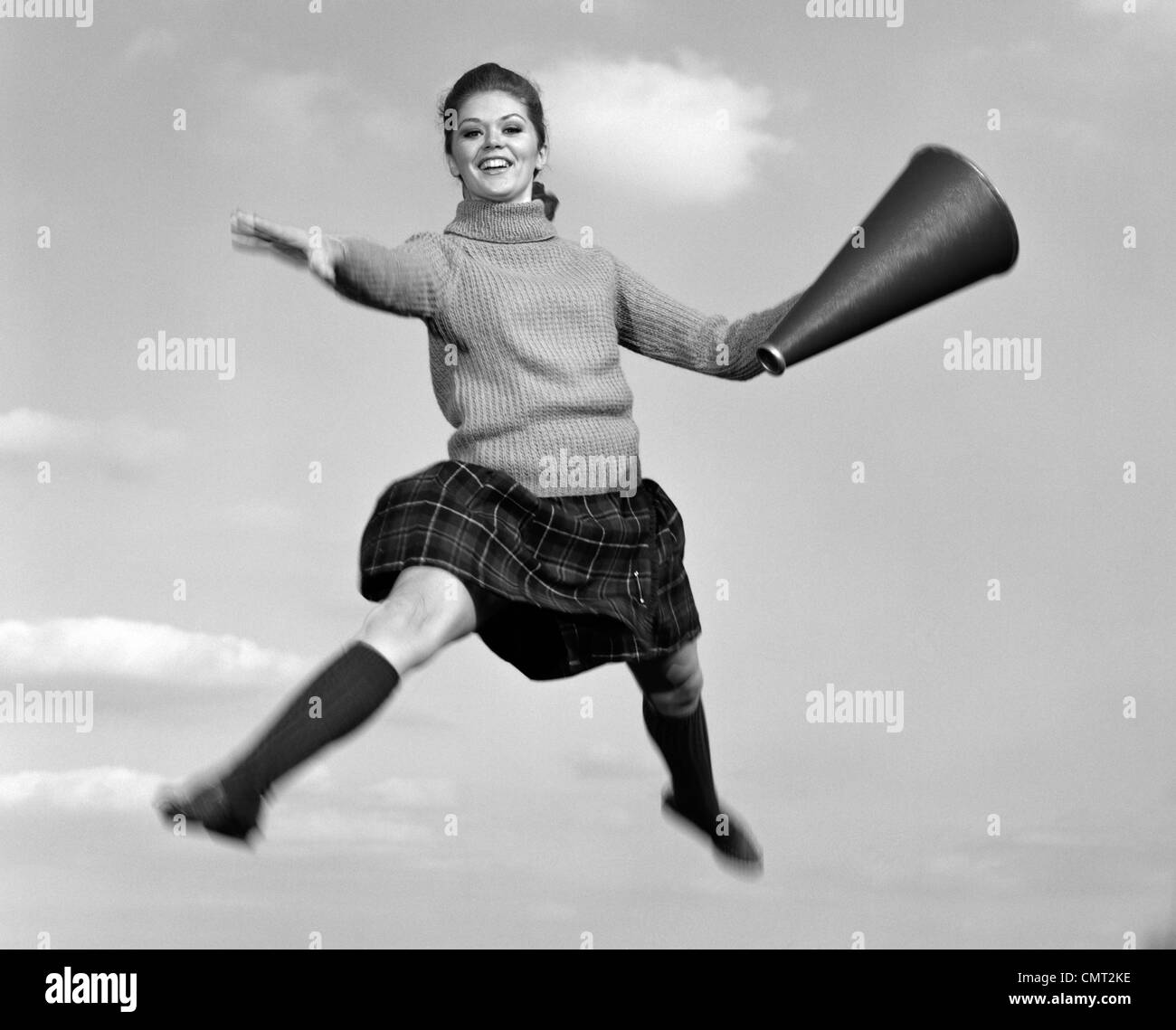 1960s TEEN GIRL CHEERLEADER JUMPING DOING SPLIT IN AIR WITH MEGAPHONE OUTDOOR LOOKING AT CAMERA Stock Photo