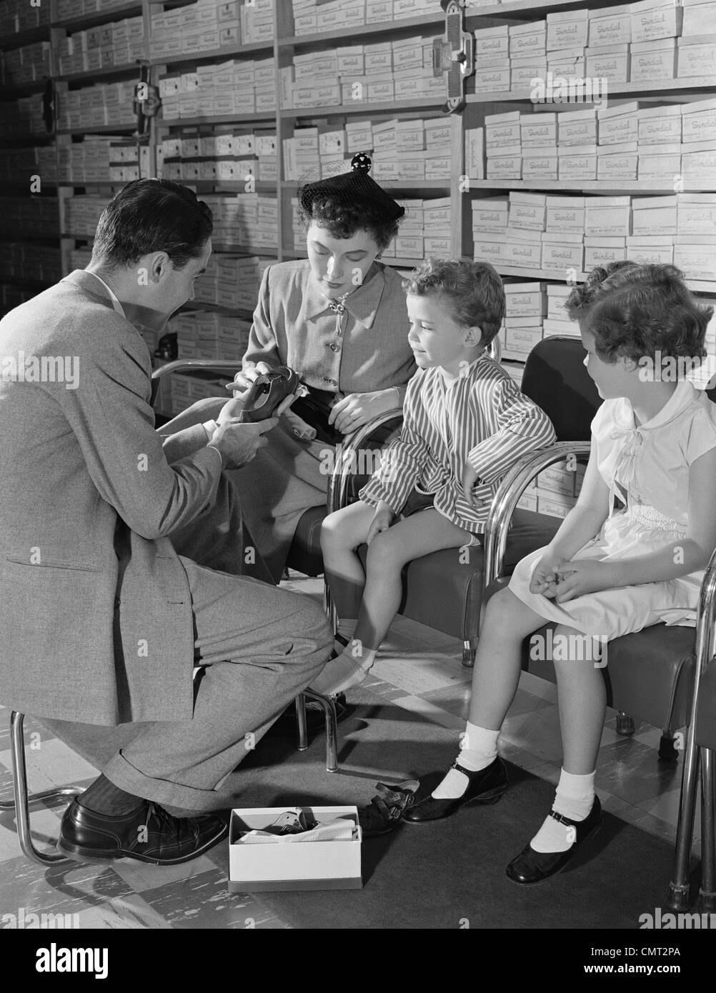 1950s FAMILY MOTHER TWO CHILDREN IN SHOE STORE TRYING NEW SHOES HELPED BY SALESMAN Stock Photo