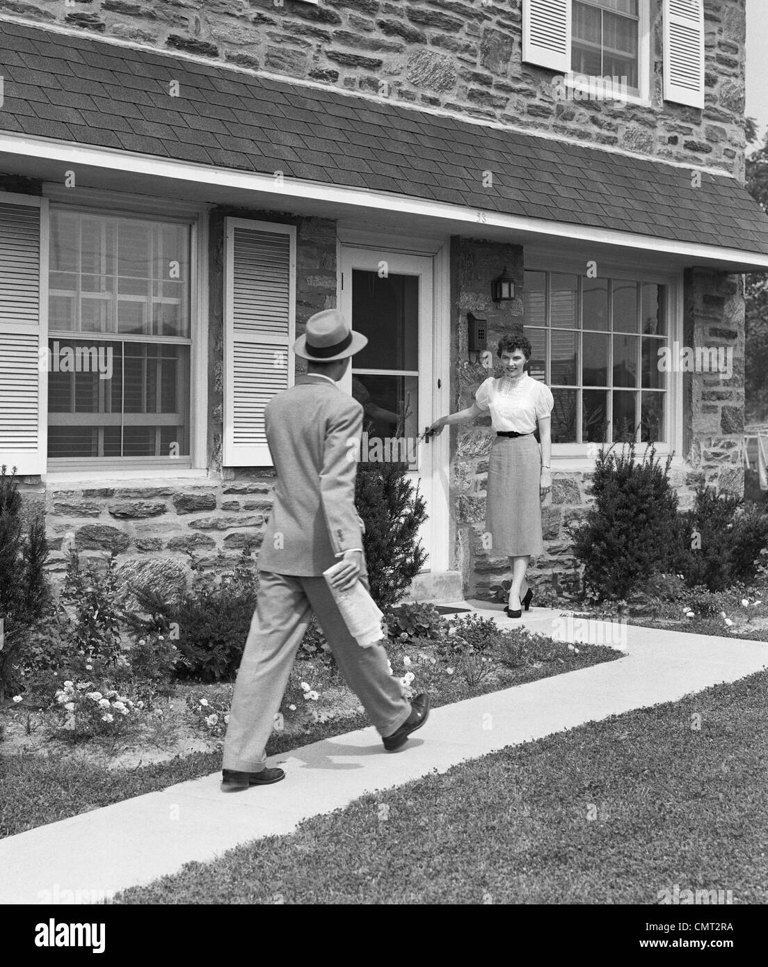 1950s MAN COMING HOME DOWN SIDEWALK WOMAN WAITING AT DOOR Stock Photo