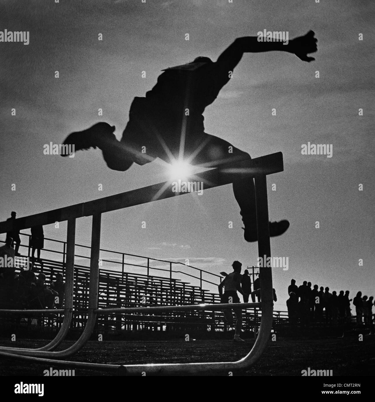 1970s SUN RAYS THROUGH SILHOUETTE OF MAN JUMPING TRACK FIELD HURDLE Stock Photo