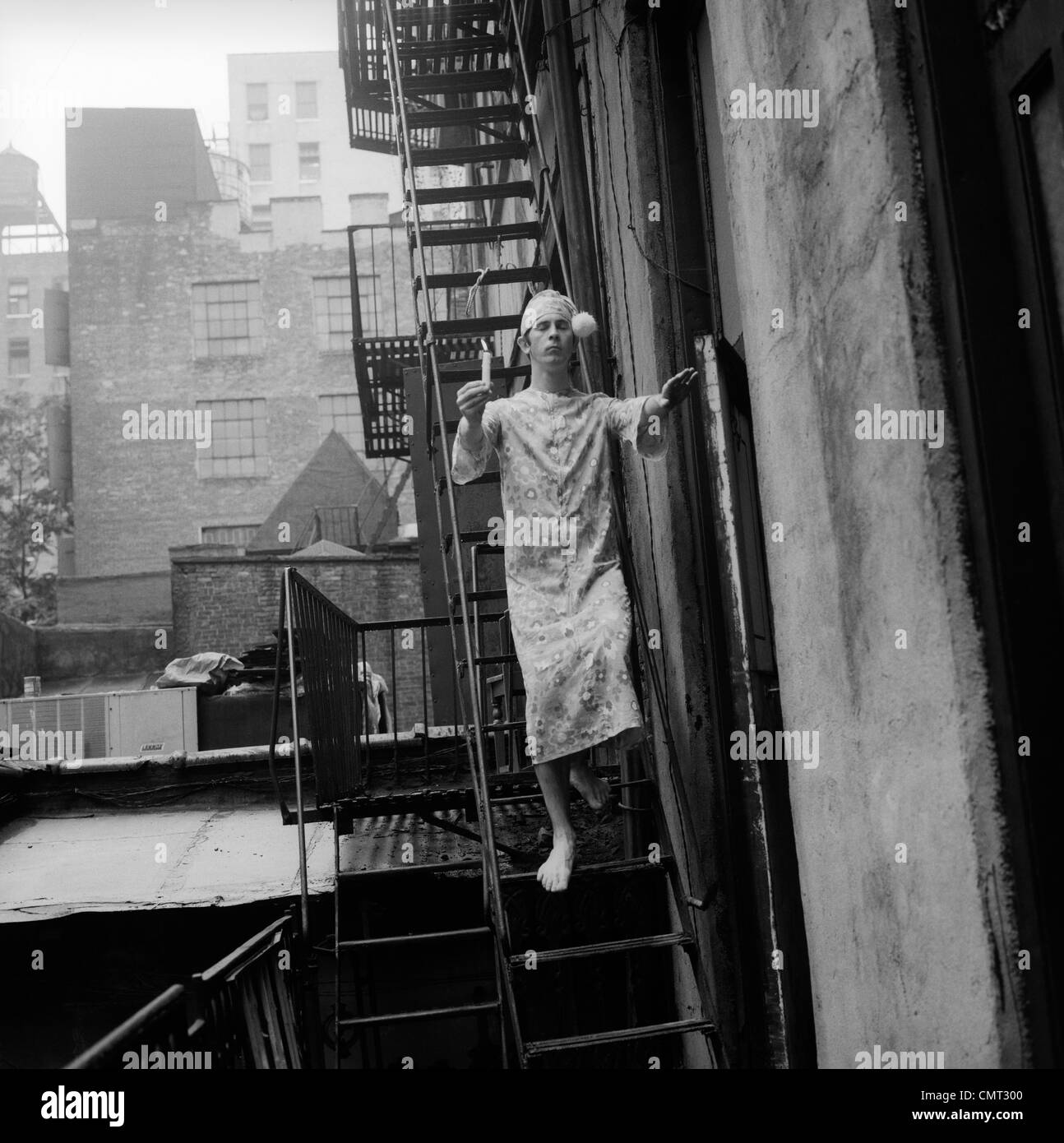 1970s MAN IN NIGHTGOWN AND CAP CARRYING CANDLE SLEEPWALKING DOWN FIRE ESCAPE OUTDOOR Stock Photo