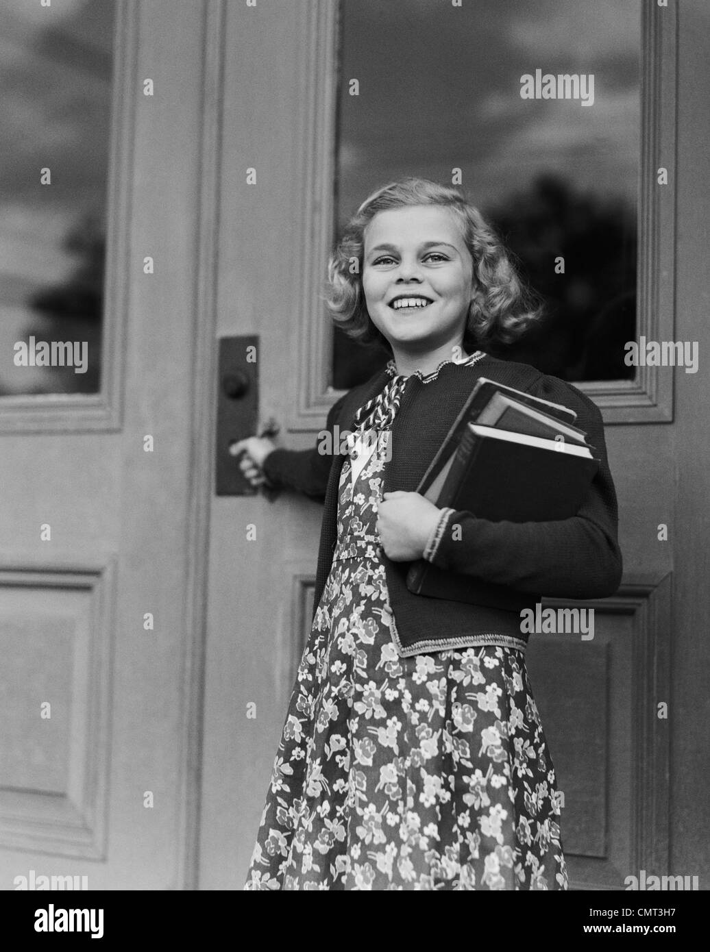 1940s SMILING GIRL PRINT DRESS HOLDING SCHOOL BOOKS OPENING DOOR Stock Photo