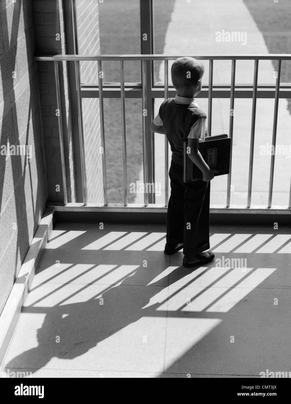1950s SCHOOLBOY WITH BOOKS UNDER ARM LOOKING OUT WINDOW BETWEEN BARS OF RAILING SHADOW CAST BEHIND HIM Stock Photo
